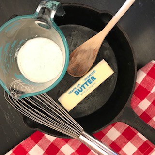 A cup of flour, a stick of butter, a whisk and a wooden spoon in a cast iron skillet.