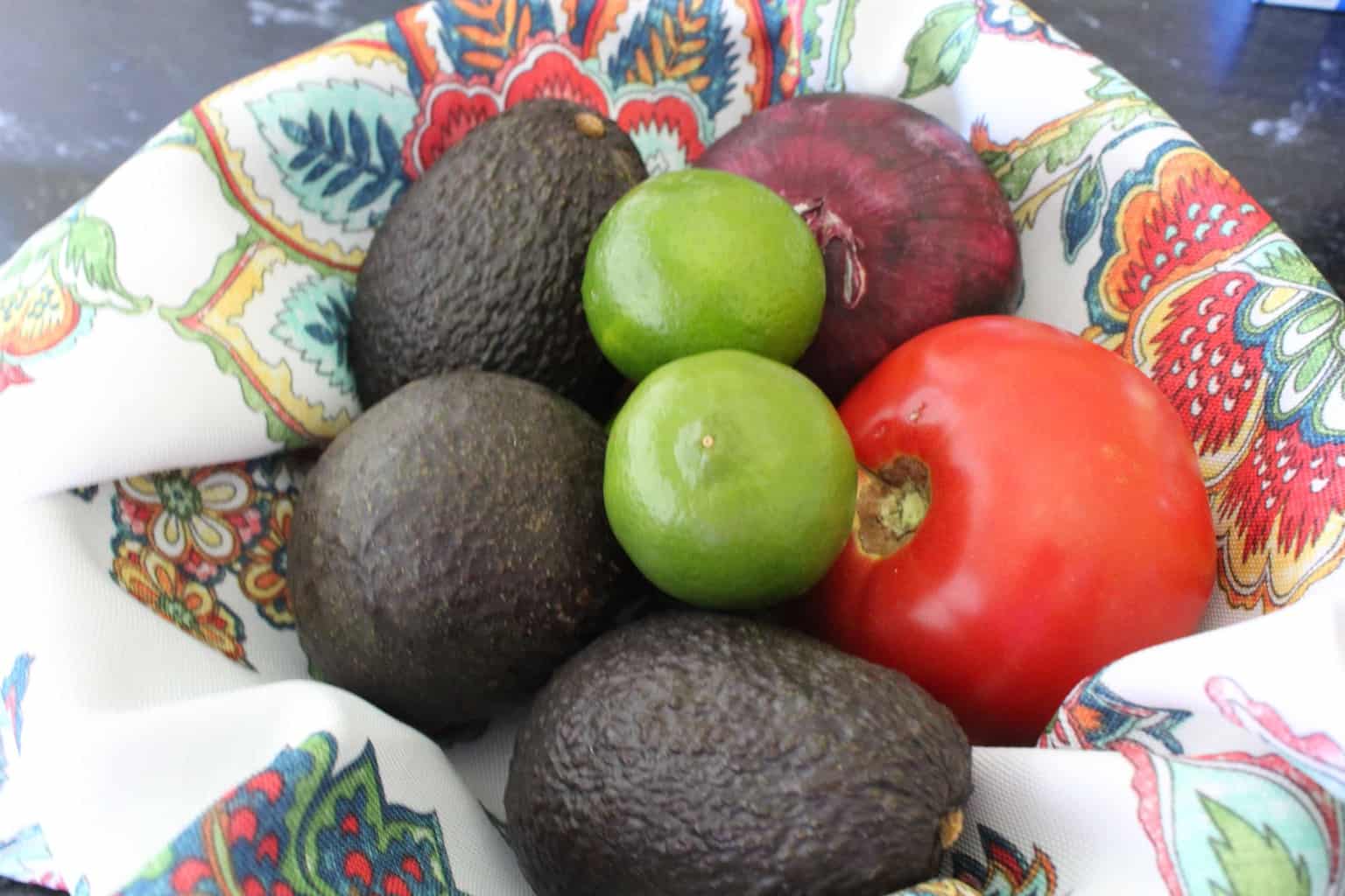 Avocados, limes, tomato and red onion in decorative bowl.