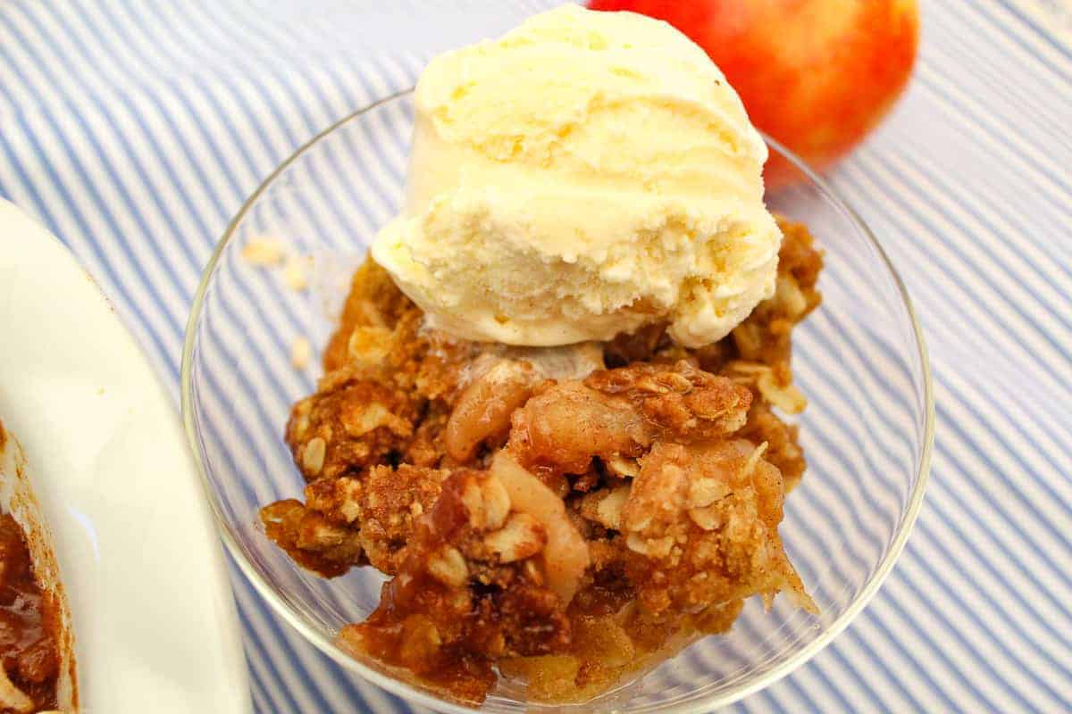 Apple crisp in clear parfait dish with a scoop of vanilla ice cream on top.