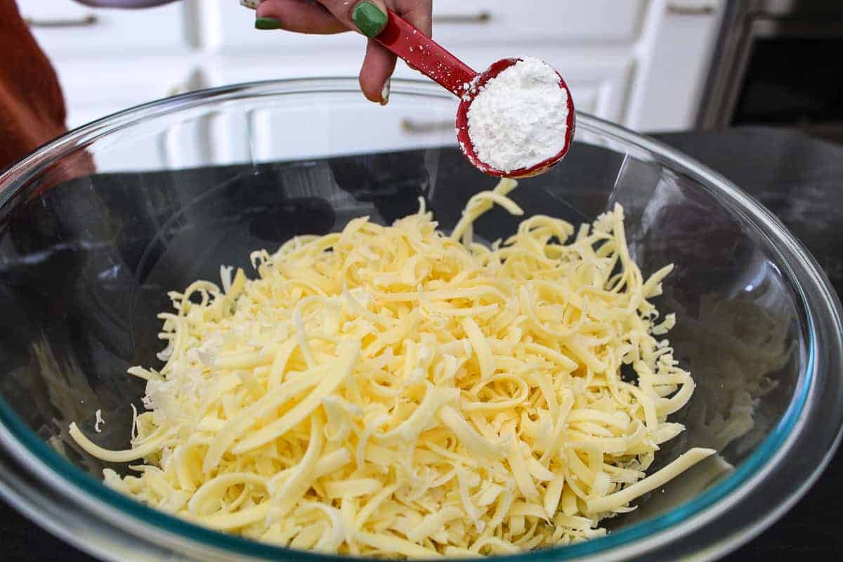 Shredded cheese in clear bowl with tablespoon of cornstarch being added