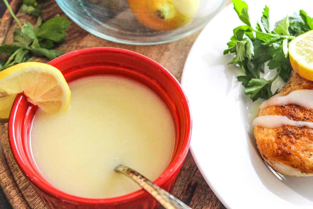 Red ramekin of lemon sauce next to plate of chicken with a drizzle of sauce