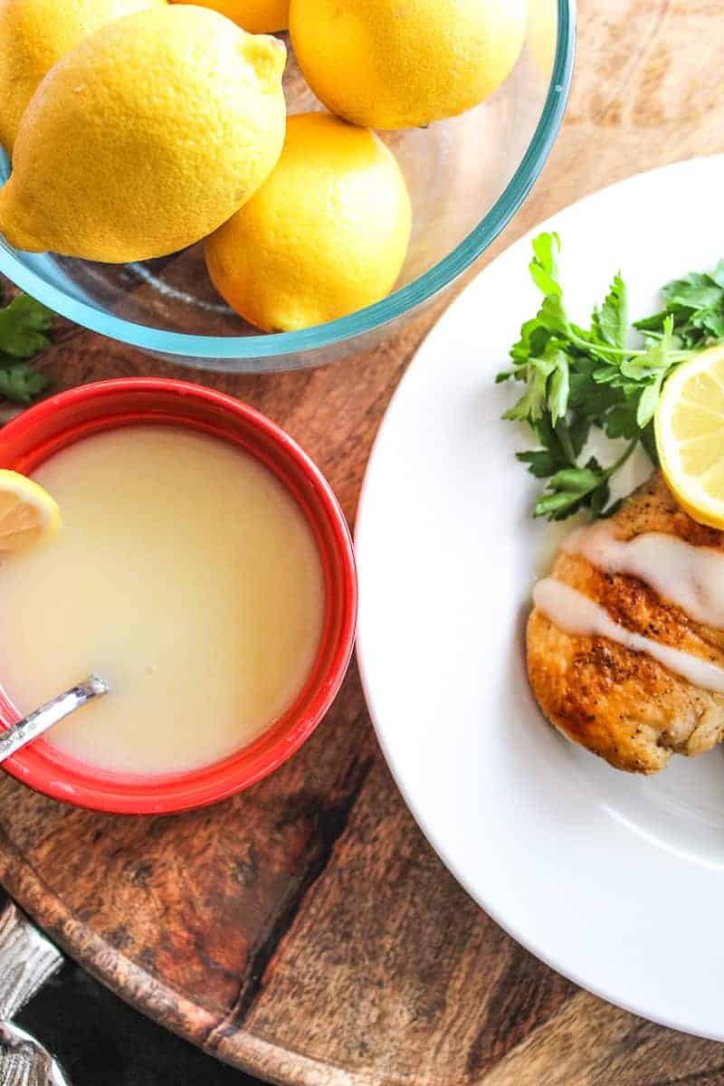 Universal Lemon Sauce in red ramekin next to plate of chicken and bowl of lemons