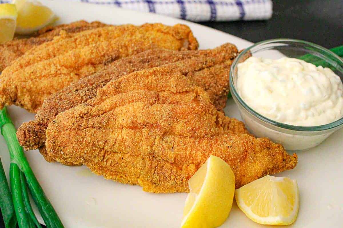 Fried catfish filets with tartar sauce in a ramekin and lemons on the side.