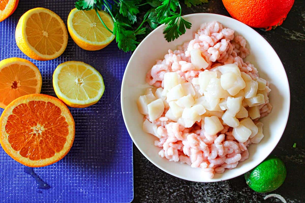 Shrimp and scallops in white bowl alongside cut lemons, limes, and orange