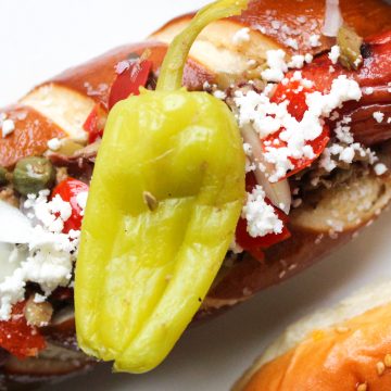 Hot dog in pretzel bun garnished with capers, feta cheese and a pepperocini