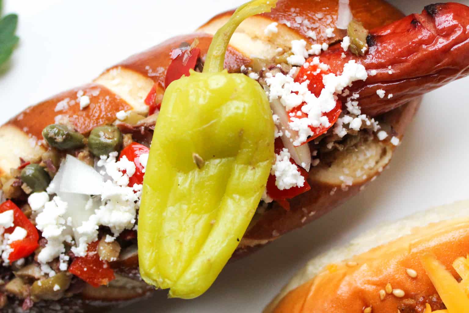 Hot dog in pretzel bun garnished with capers, feta cheese and a pepperocini