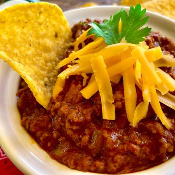 Authentic Texas Chili in white bowl with tortilla chip and shredded cheese.