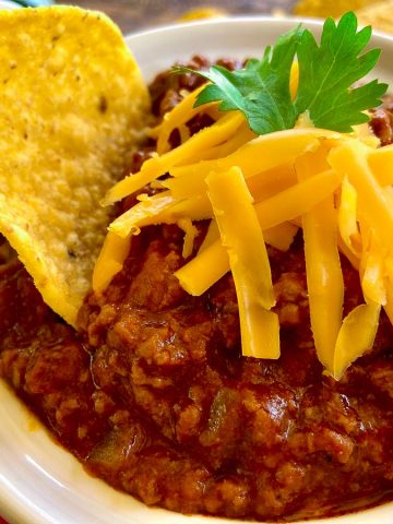 Authentic Texas Chili in white bowl with tortilla chip and shredded cheese.