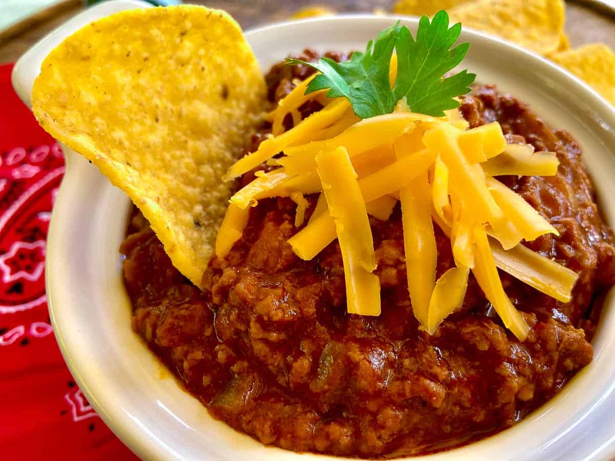 Authentic Texas Chili in white bowl with tortilla chip and shredded cheese.