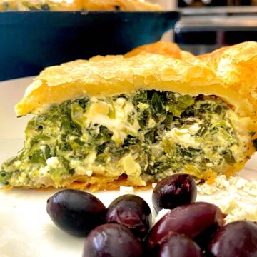 Slice of spinach feta pie on white plate with kalamata olives and feta crumbles