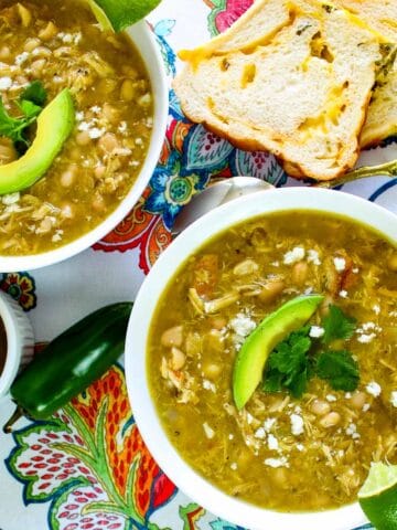 Two white bowls of green chicken chile on floral tablecloth with side of jalapeno