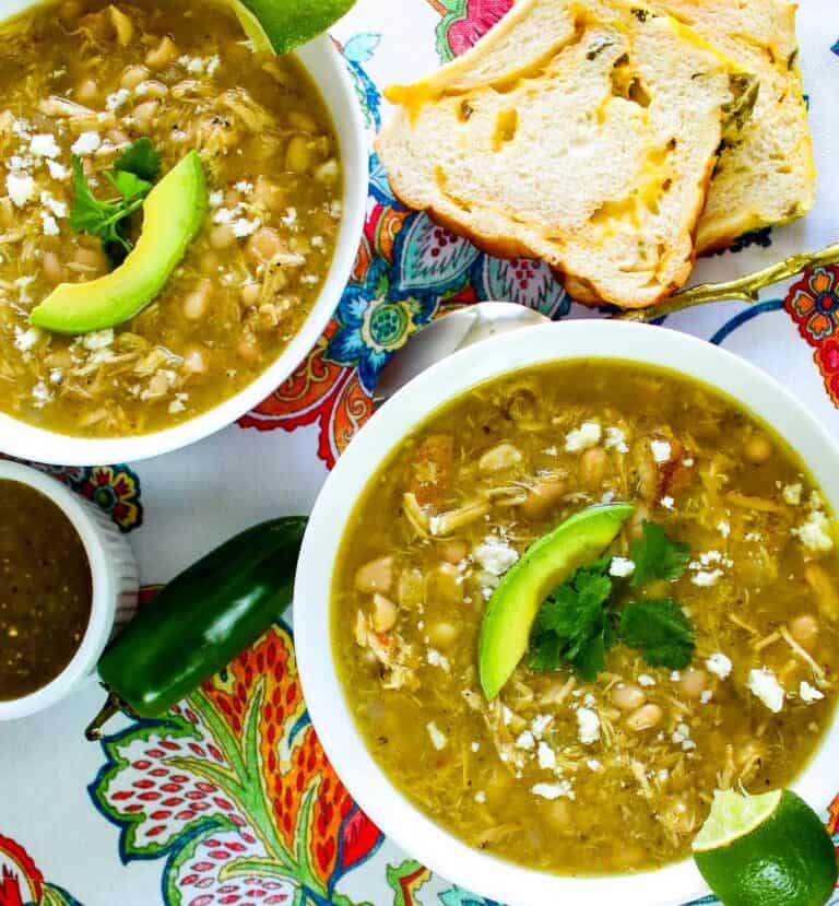 Two white bowls of green chicken chile on floral tablecloth with side of jalapeno