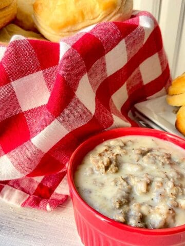 Sausage cream gravy in red bowl with bowl of biscuits in red checkered basket in background