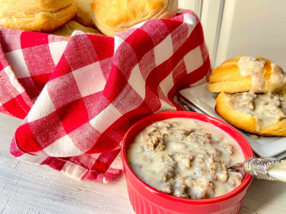 Sausage cream gravy in red bowl with bowl of biscuits in red checkered basket in background