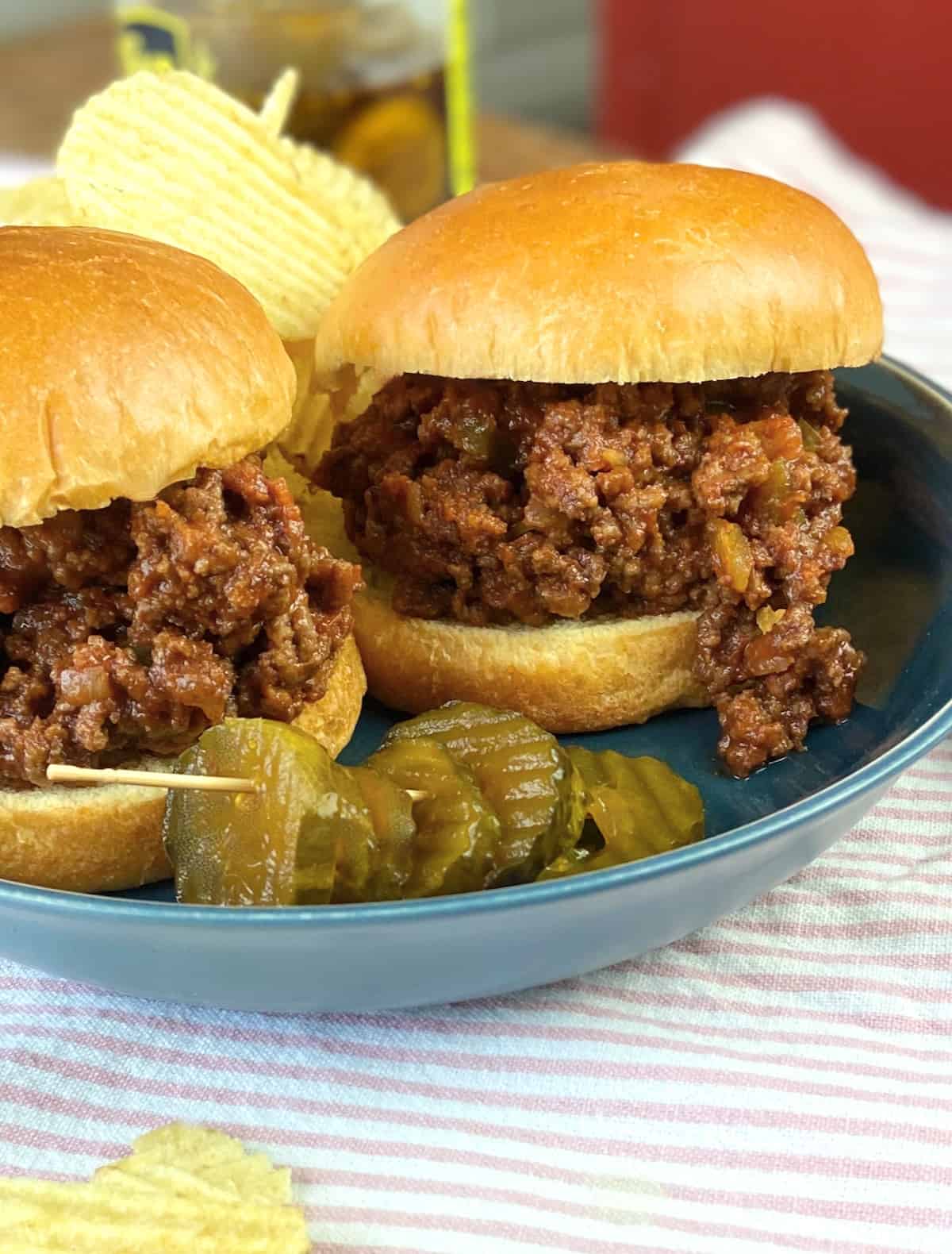 Two sloppy joe's sandwiches on blue plate with chips and pickles.