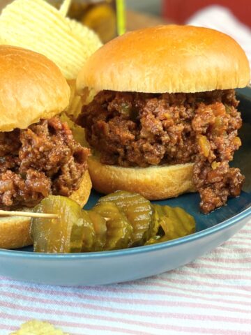 Two sloppy joe's on a blue plate with pickles.