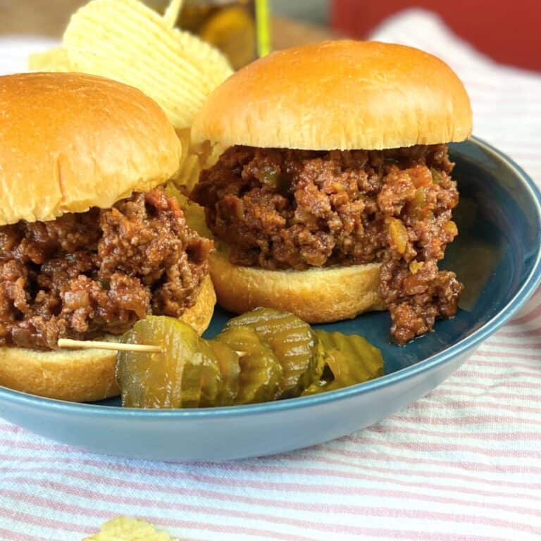 Two sloppy joe's on a blue plate with pickles.