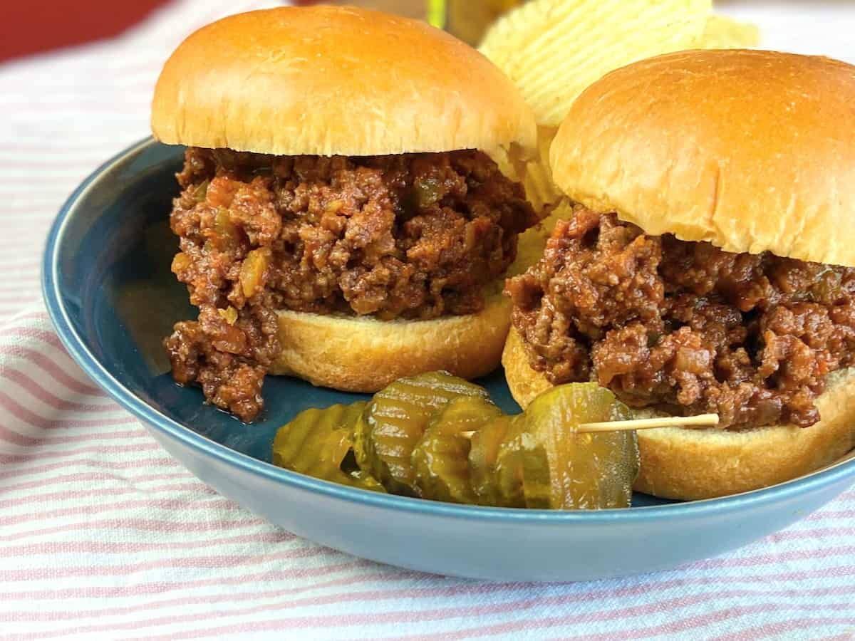 Two sloppy joe's on a blue plate with chips and pickles.