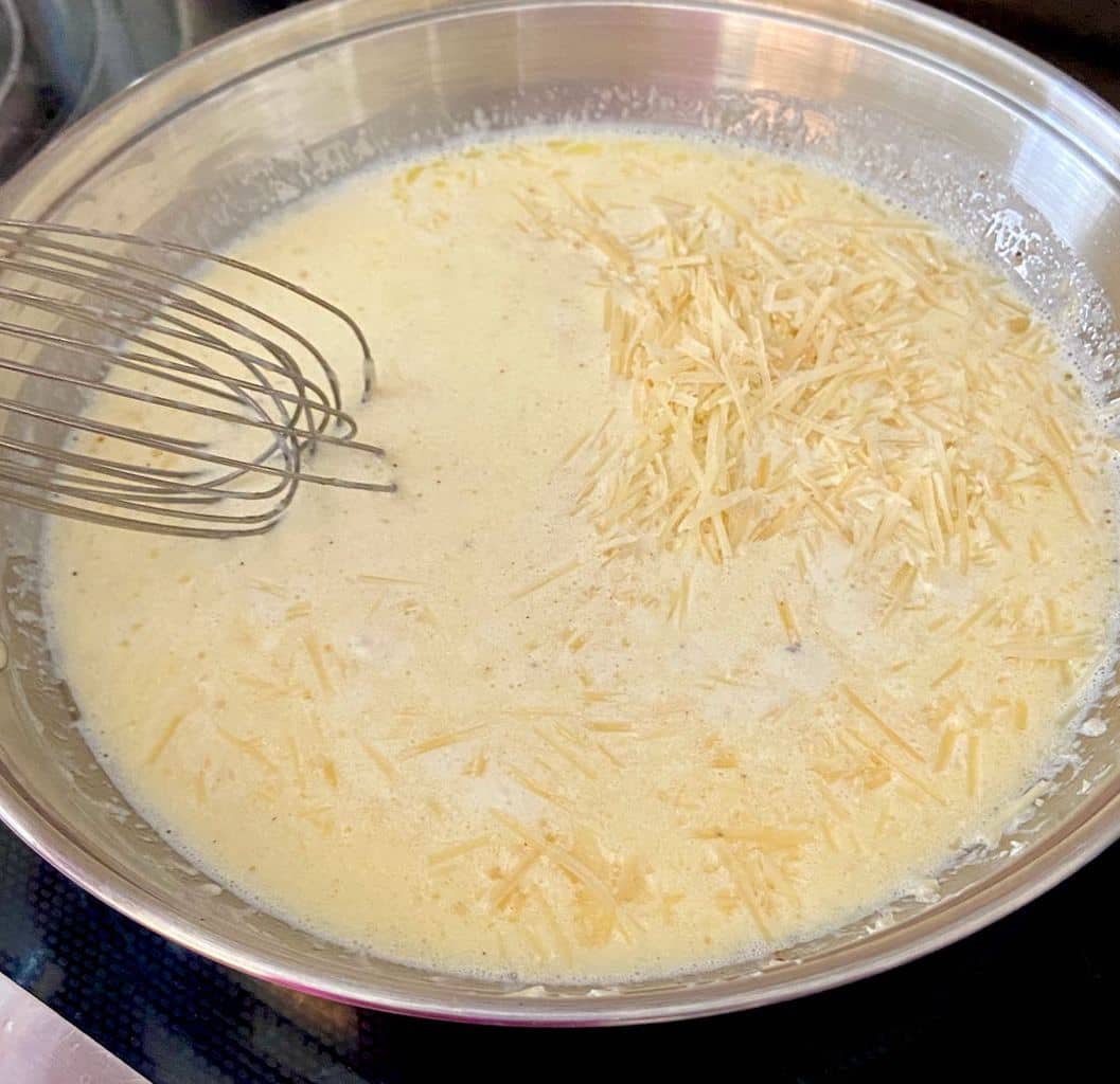 Alfredo sauce with shredded parmesan in aluminum skillet being whisked with a wire whisk.