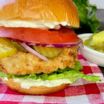 Fried chicken sandwich with all the fixins on red and white plaid tablecloth.