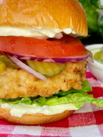 Fried chicken sandwich with all the fixins on red and white plaid tablecloth.