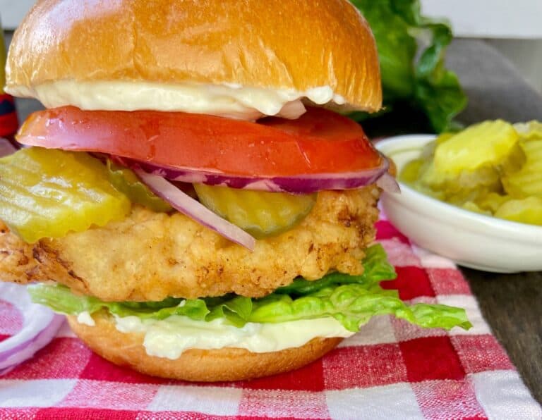 Fried chicken sandwich with all the fixins on red and white plaid tablecloth.
