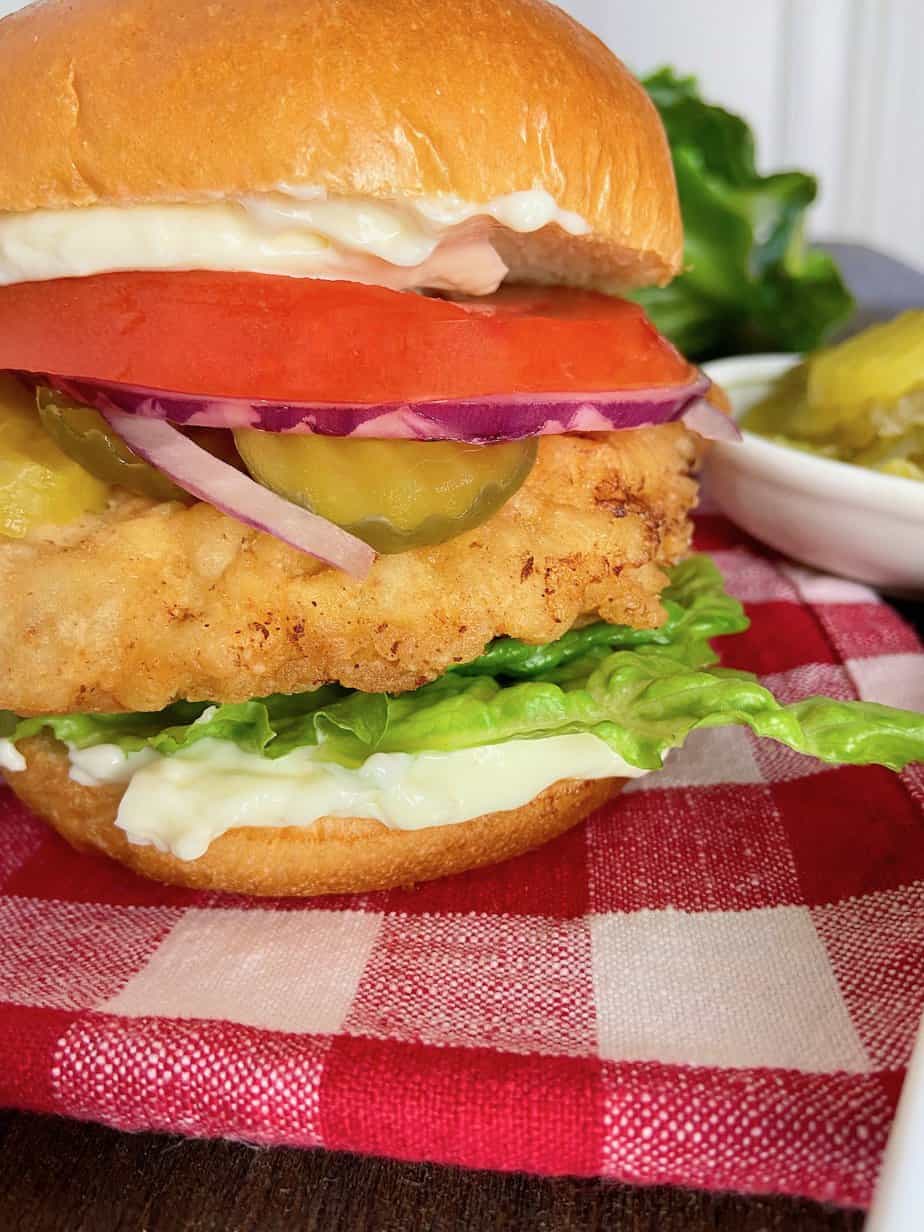 Fried Chicken Sandwich with lettuce, tomatoes, onion, dill pickles and mayonnaise on red and white plaid tablecloth.