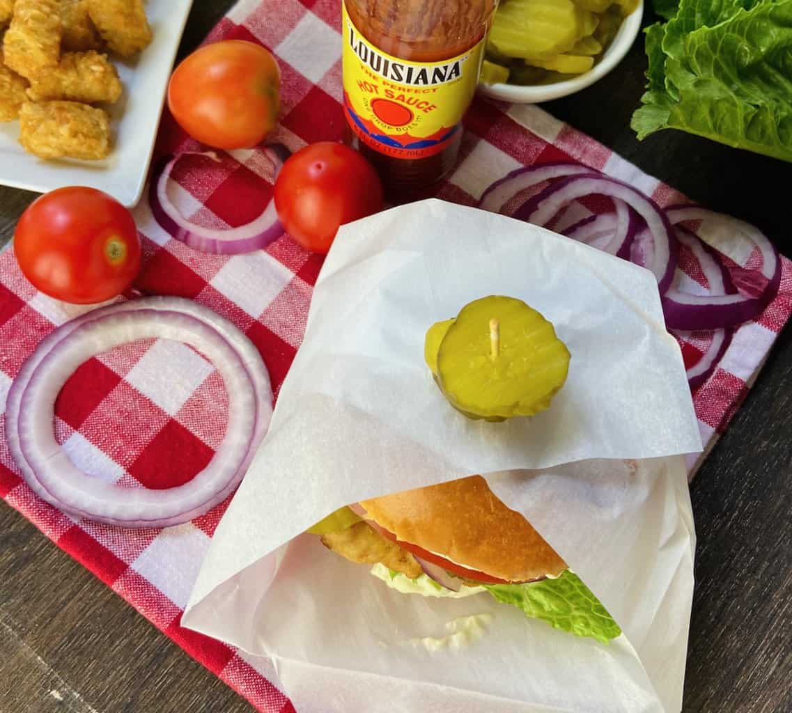 Sandwich in paper on red and white picnic table with topping on the table.