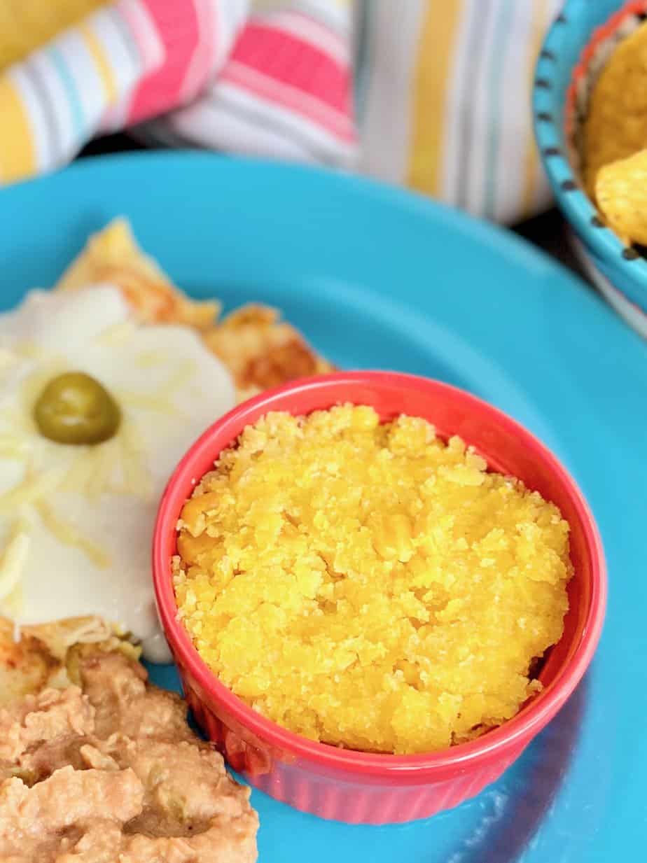 A red ramekin of sweet corn tomalito on a blue plate with enchiladas and refried beans.