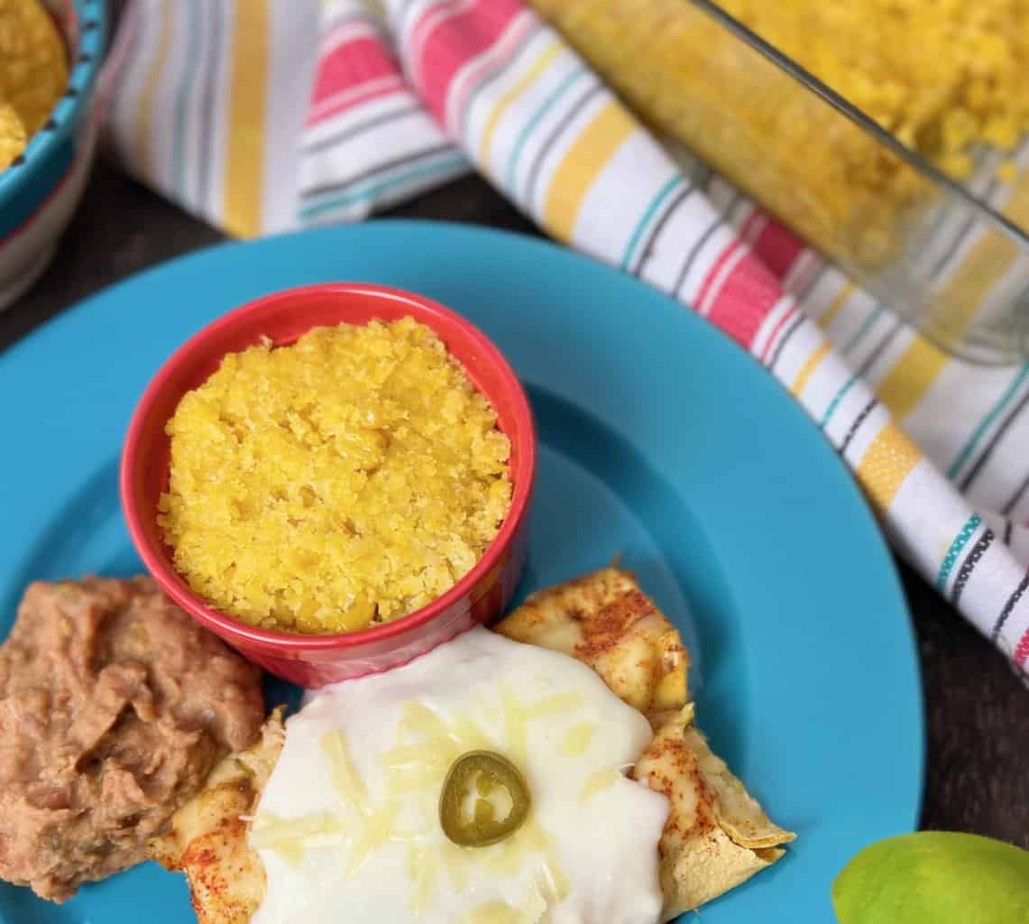 A red ramekin of sweet corn tomalito on a blue plate with enchiladas and refried beans.