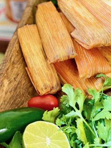 Tamales in corn husks stacked on wooden platter.