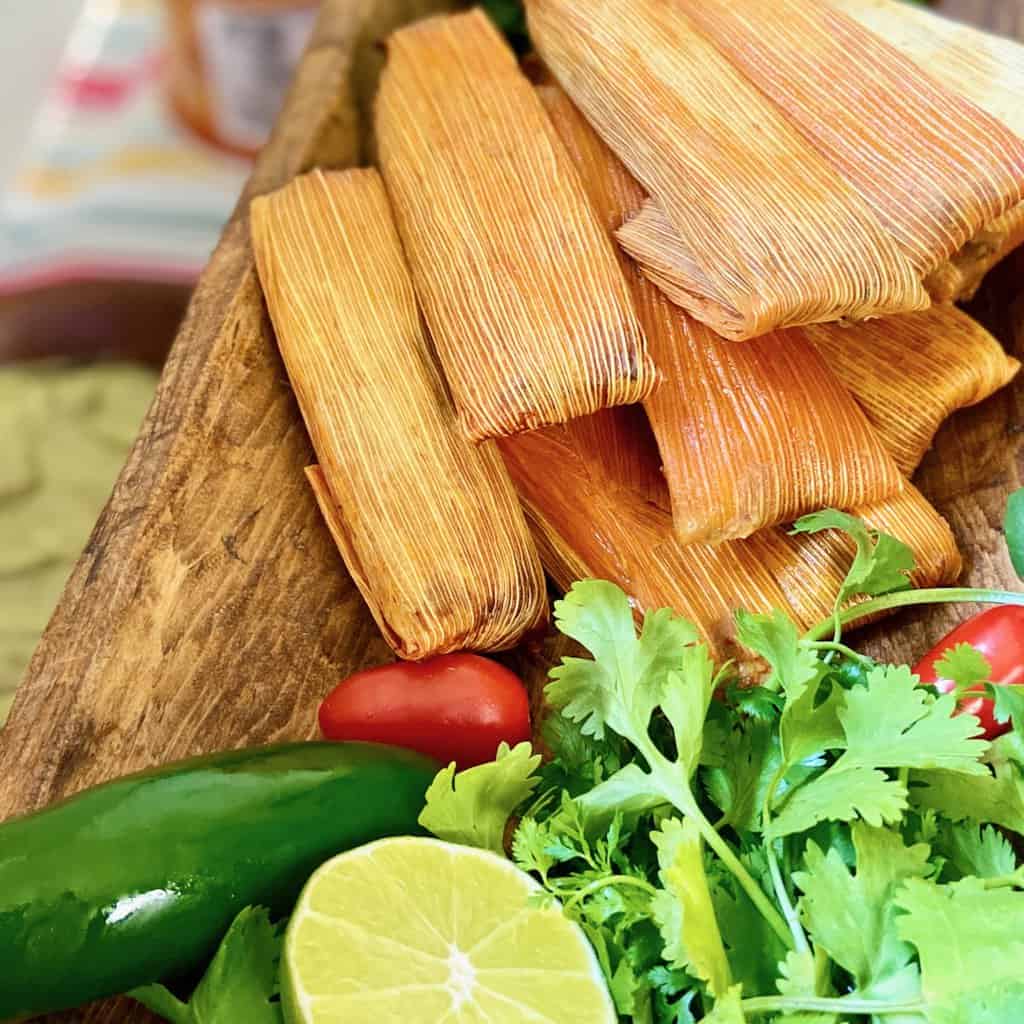 Tamales in corn husks stacked on wooden platter.