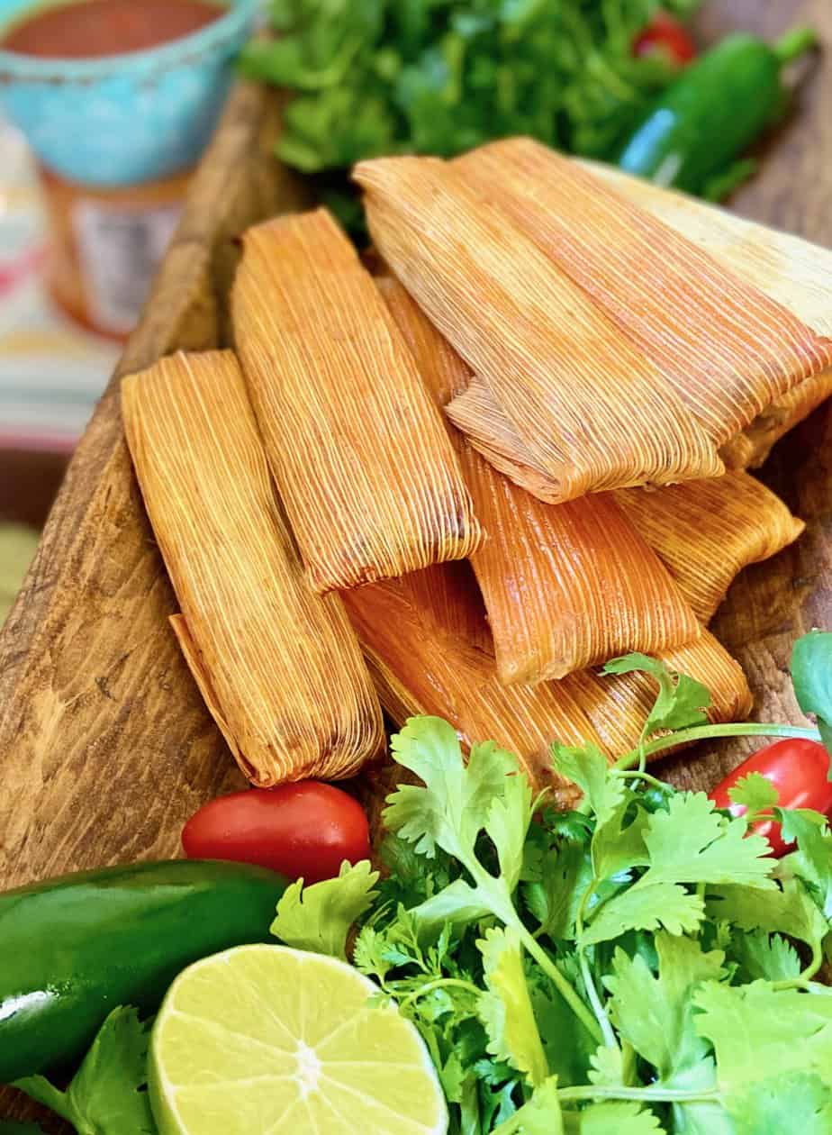 Tamales stacked on wooden platter garnished with cilantro and lime.