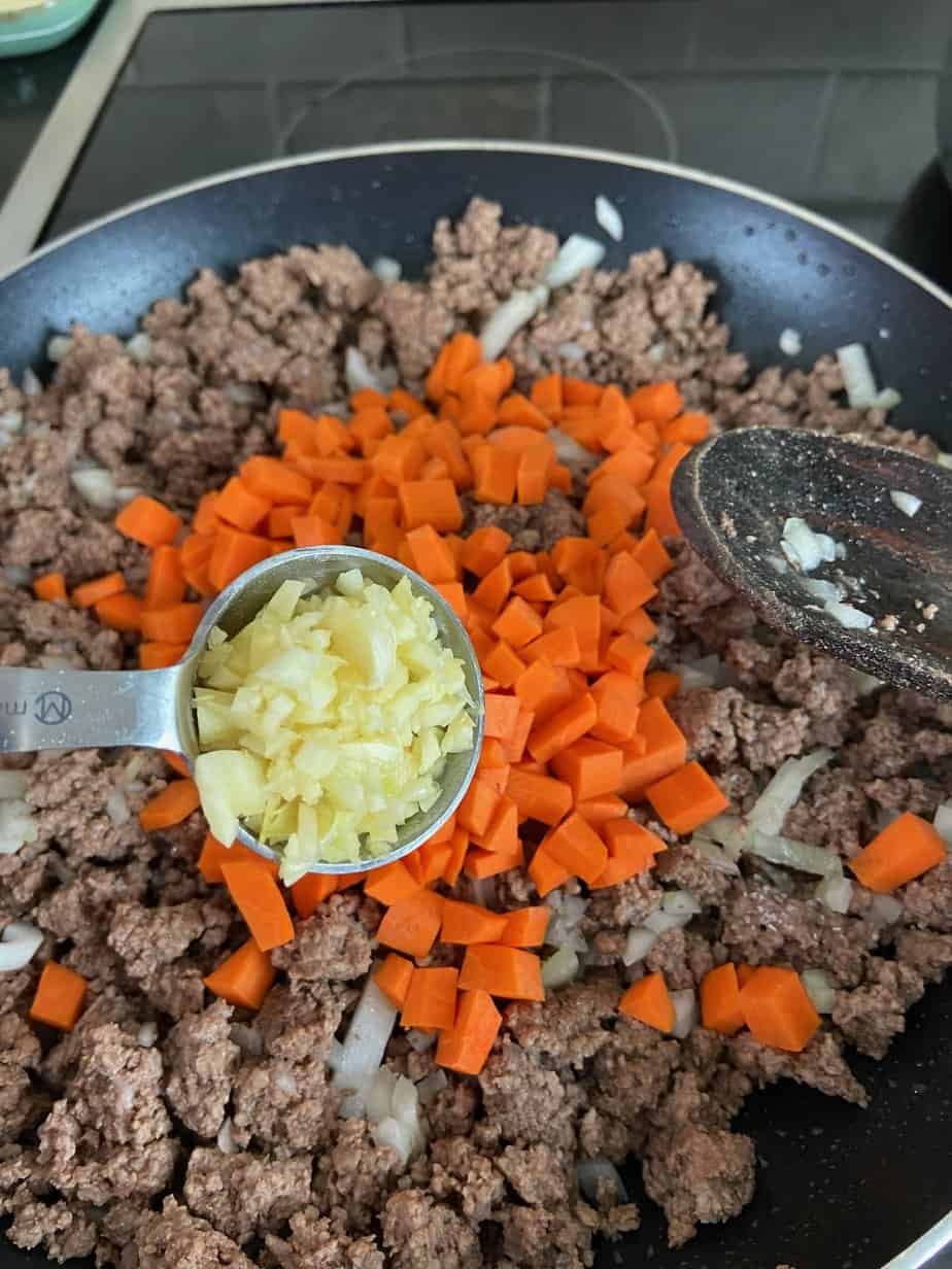 Minced garlic being added to browned beef, onions and carrots in skillet.