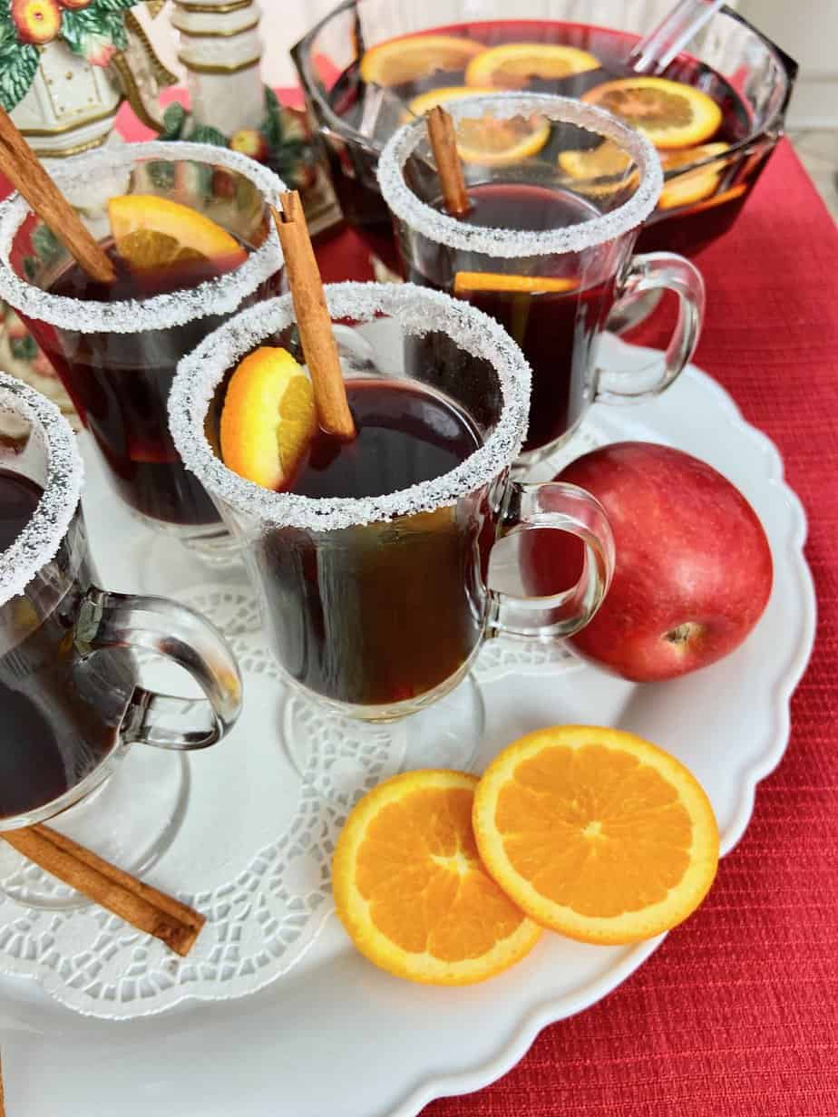Glass mugs of mulled red wine on white platter garnished with orange slices.