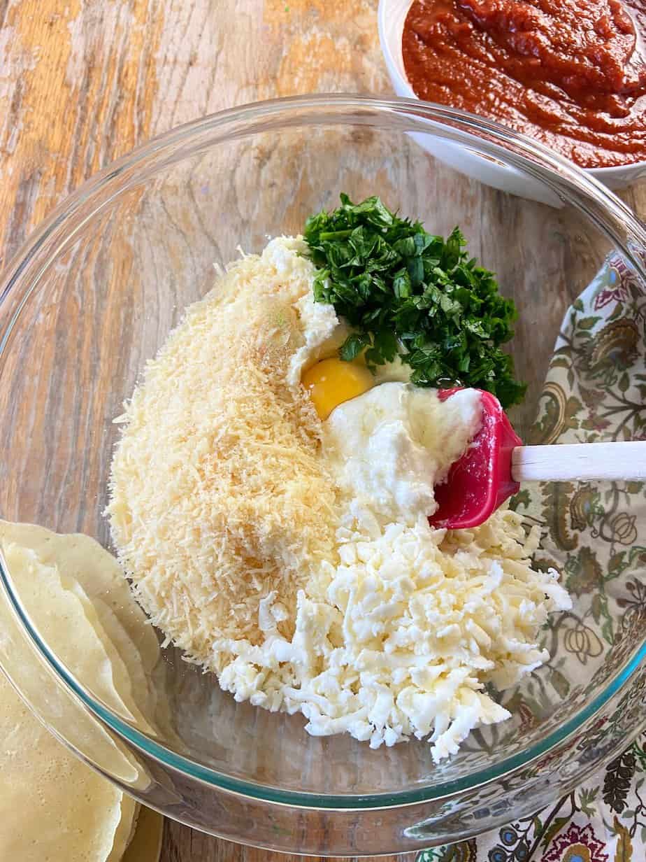Ricotta, parmesan, mozzarella cheese, fresh parsley and one egg in glass mixing bowl.