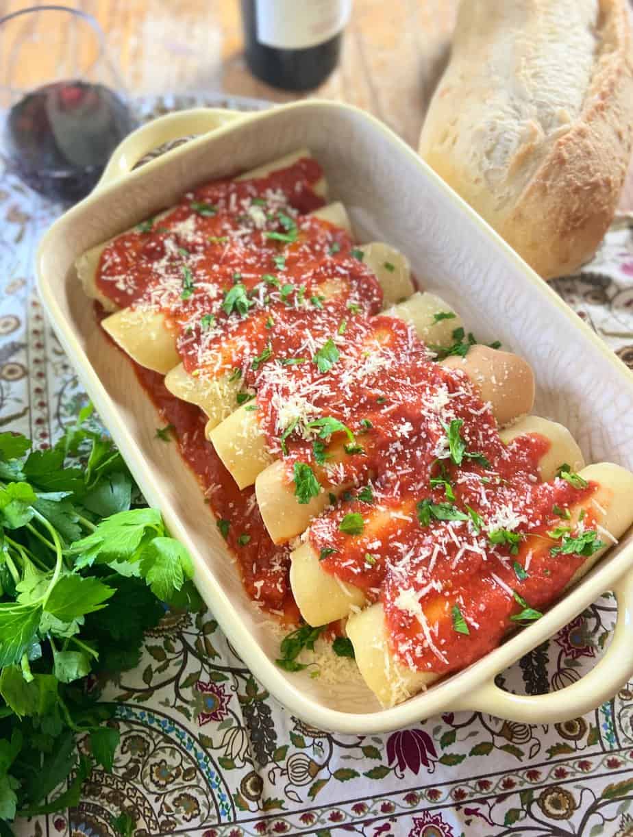 Manicotti crepes in a yellow baking dish with marinara sauce, grated parmesan cheese and fresh parsley on top.