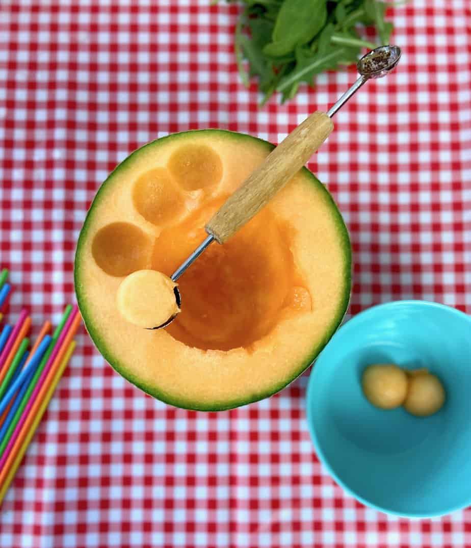 Melon baller scooping out cantaloupe on checkered tablecloth.