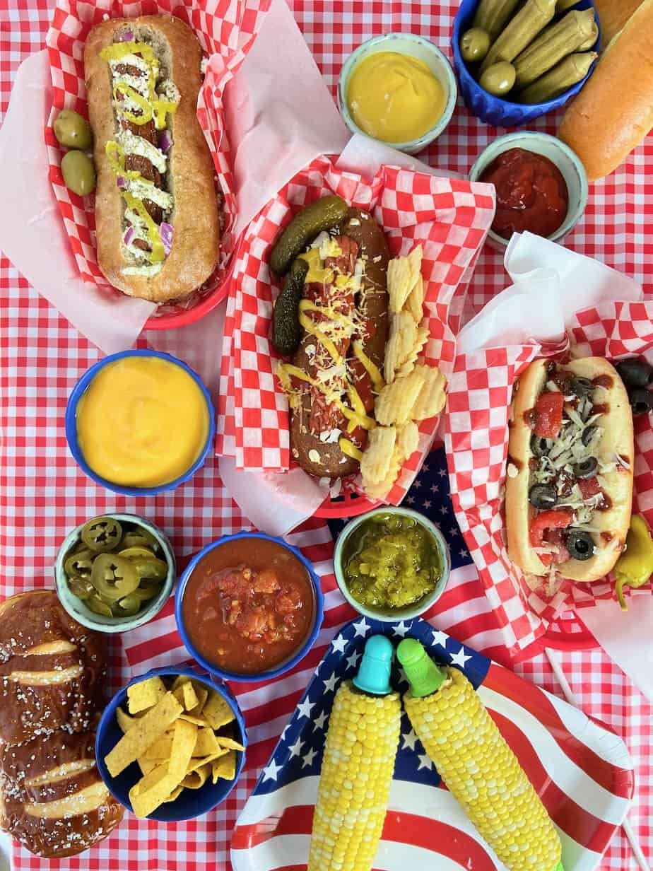 Hot dog bar toppings, sauces and sides on red checkered tablecloth.