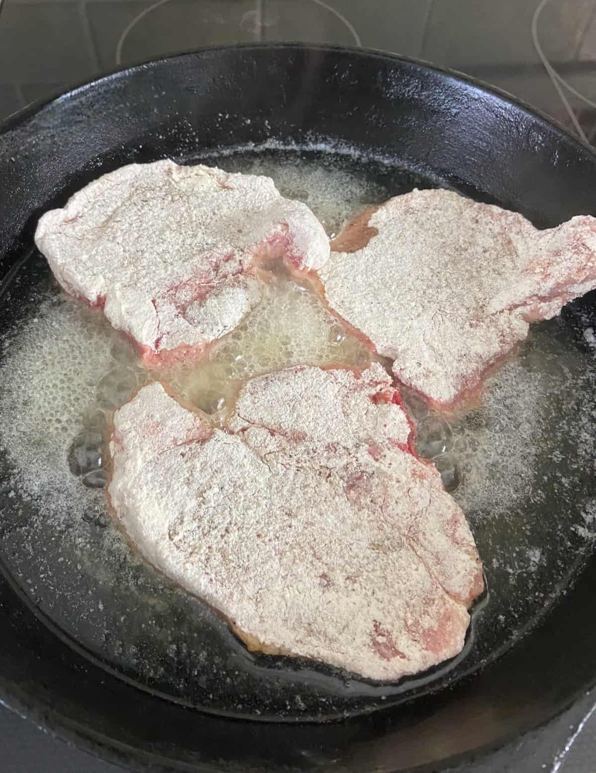 Three floured pork chops frying in cast iron skillet in oil.