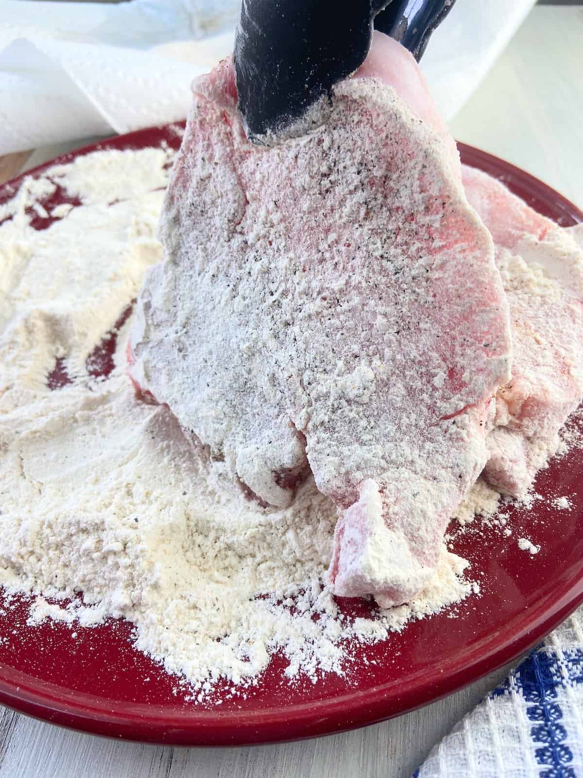 Floured pork chop being held over flour mixture with tongs.