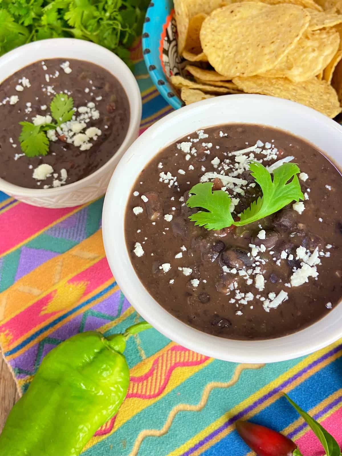 Two bowls of black bean soup with a side of tortilla chips on a colorful tablecloth.