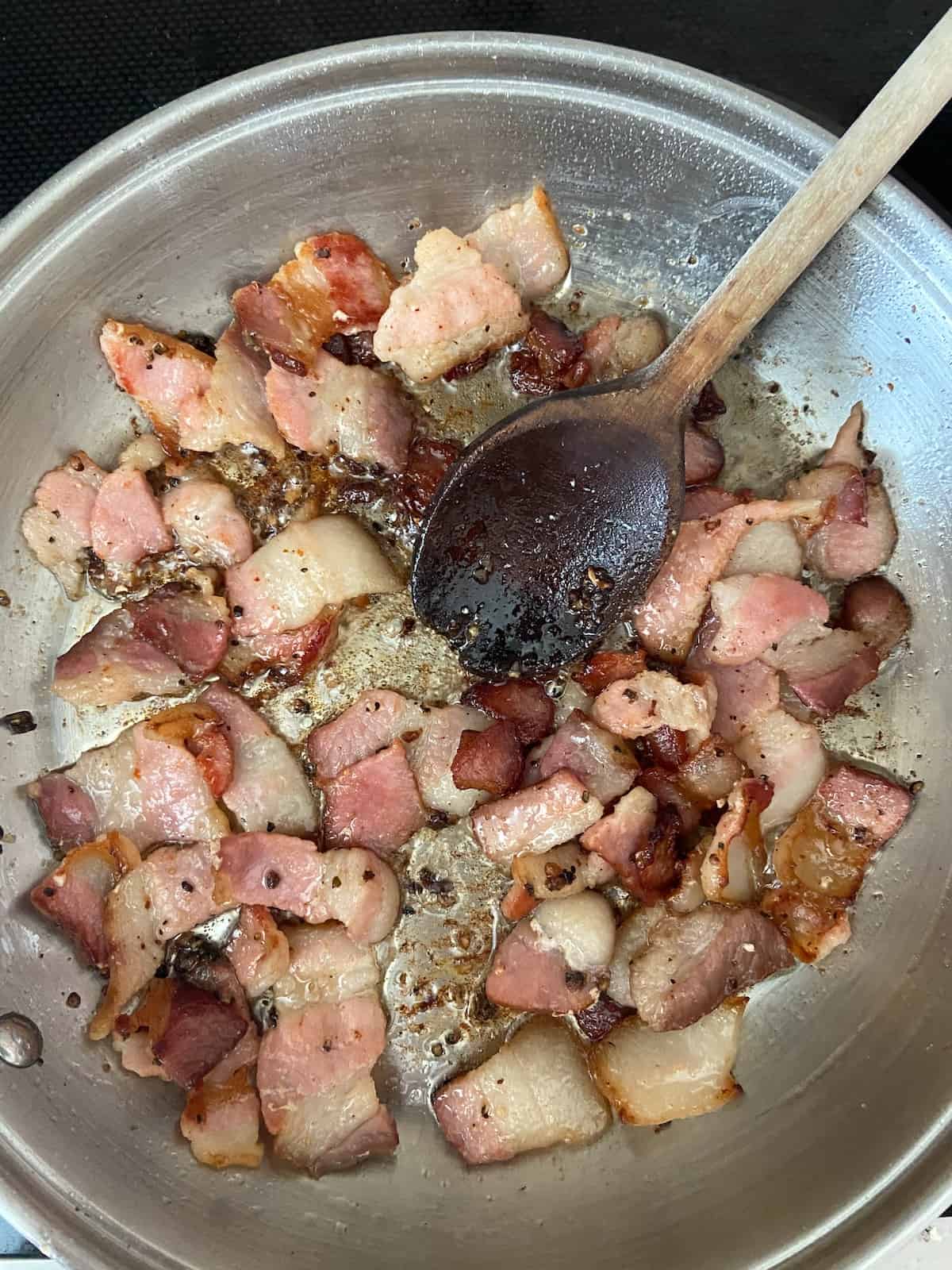 Bacon pieces in frying pan with a wooden spoon.