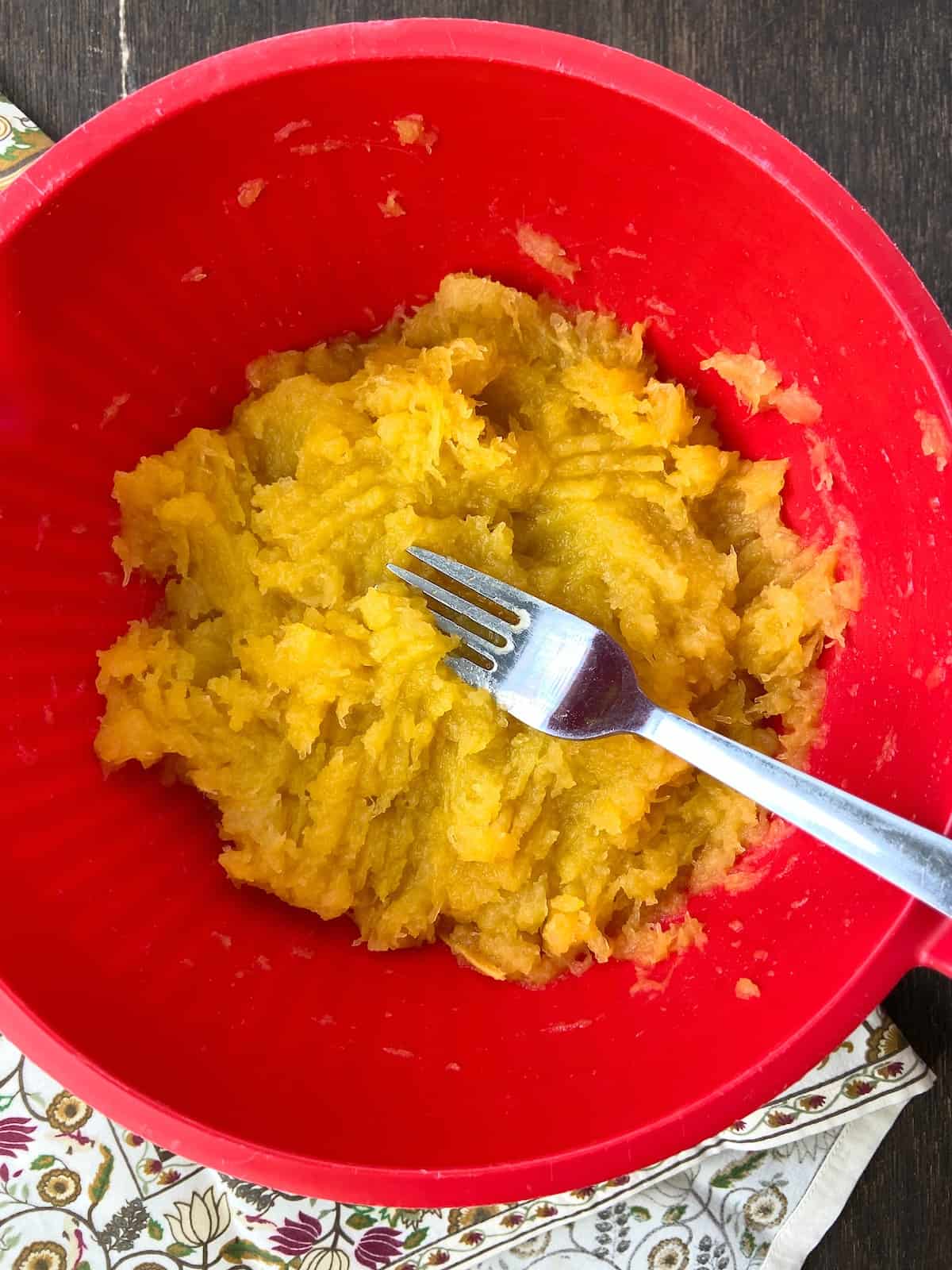 Cooked acorn squash in red bowl being mashed with fork.