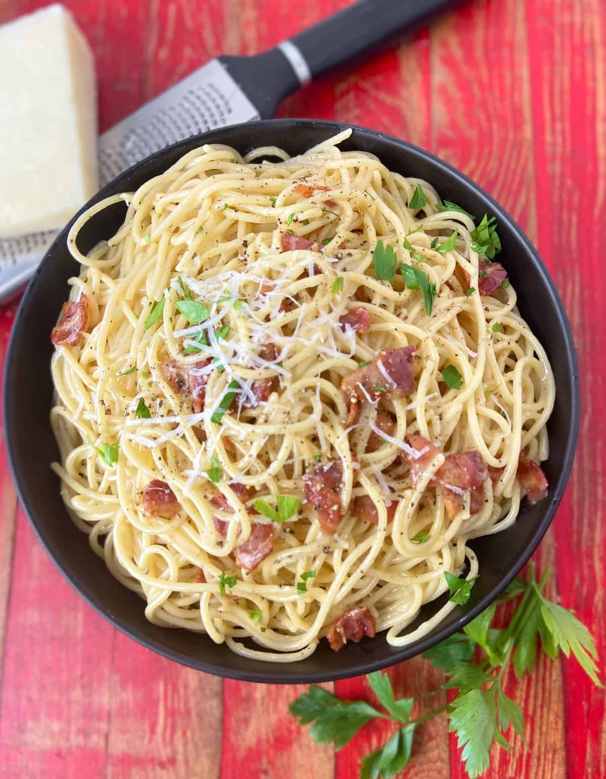 Pasta Carbonara in black bowl garnished with grated cheese and parsley and bacon bits.
