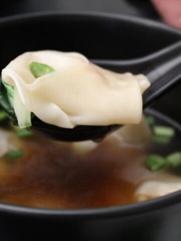 Wonton on black spoon over a bowl of wonton soup.