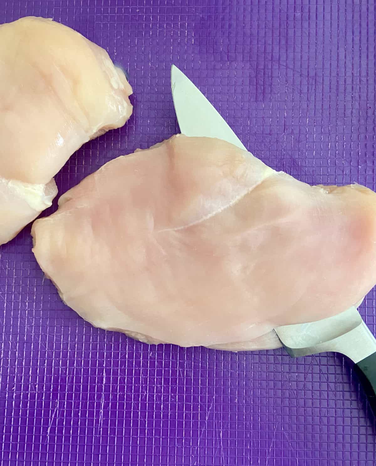 Chef's knife slicing chicken breast horizontally on purple cutting board.