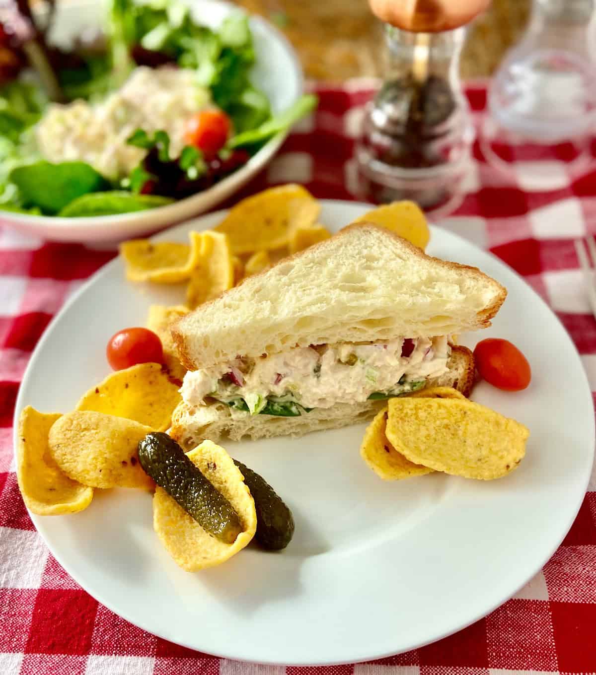 Tuna salad sandwich on white plate with corn chips and a pickle.