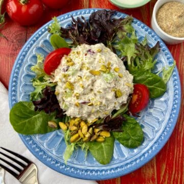 Pistachio chicken salad on a bed of greens on a blue plate with tomato wedges.