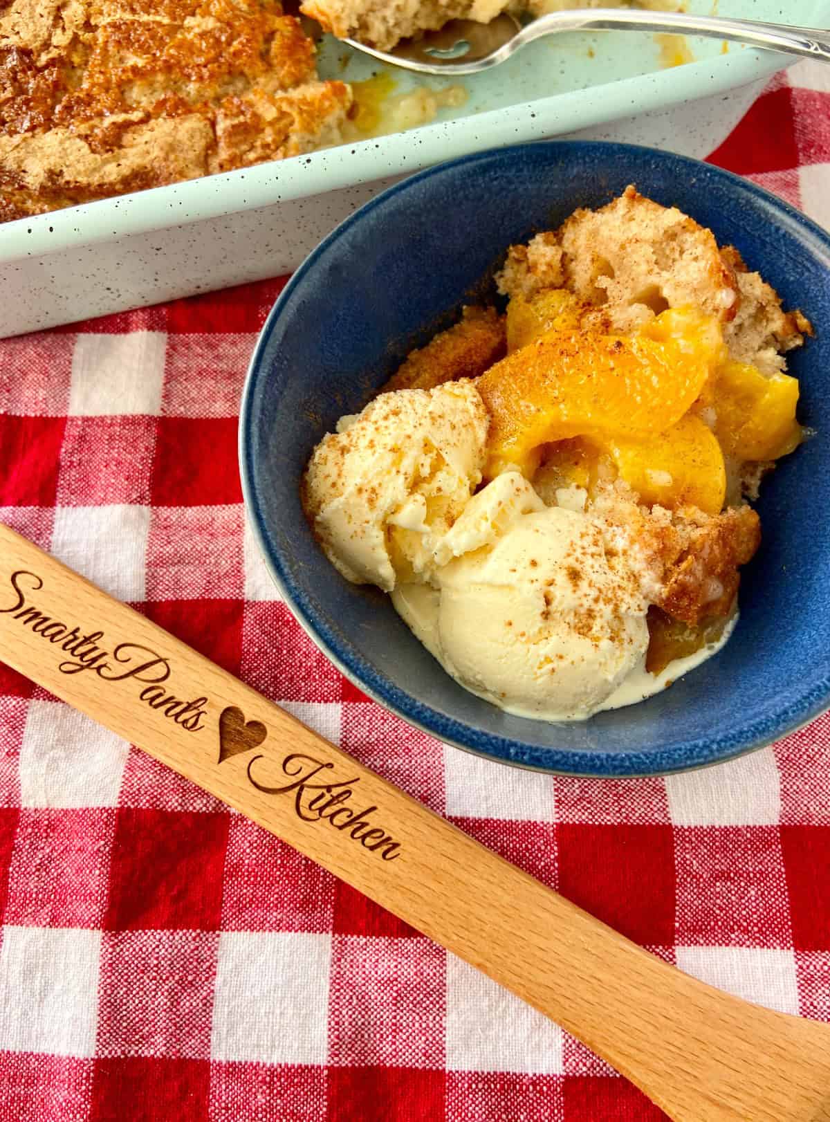 Peach cobbler in blue bowl on red and white checkered tablecloth.